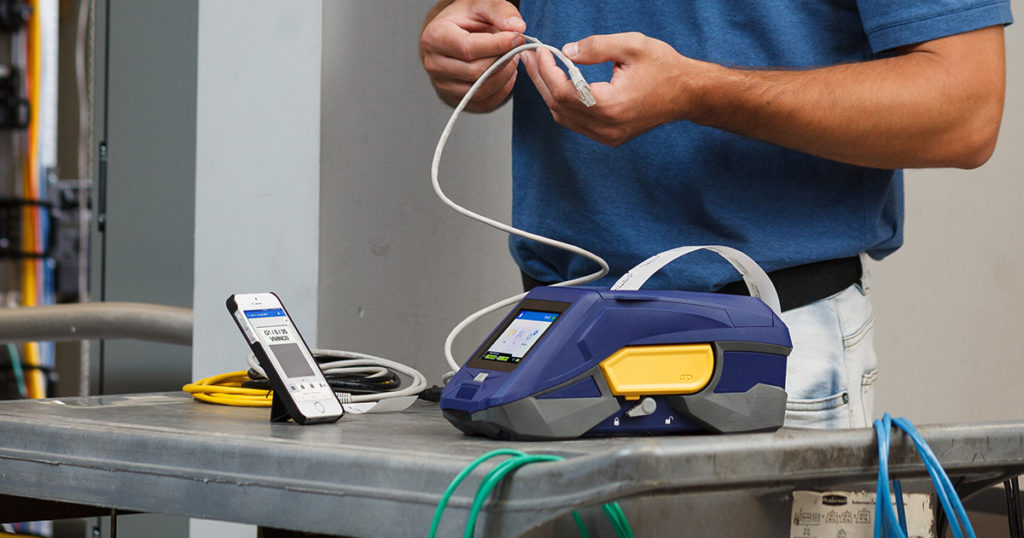 A portable label printer and connected smartphone app sit on a work cart while a technician applies labels to a cable.