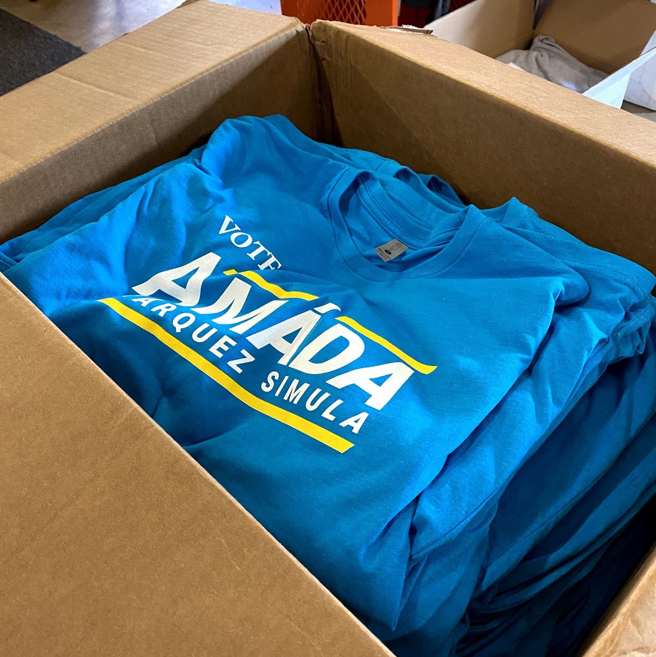 A cardboard box full of blue T-shirts printed Vote Amáda Márquez Simula.
